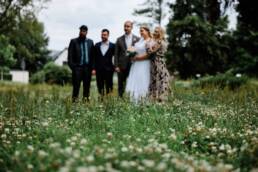 Gruppenaufnahme der Braut mit den Trauzeugen, das Gras im Vordergrund steht im Fokus. Sie lachen alle Richtung Kamera.