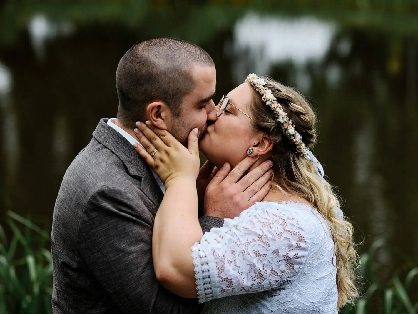 Intimer Kuss des Brautpaares im Park, Hände halten liebevoll die Gesichter fest. Idyllischer Teich im Hintergrund.
