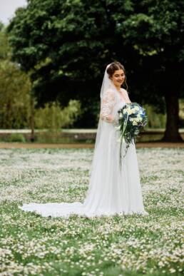Die Braut steht auf dem traumhaften Gänseblümchen Feld, sie hält ihren Hochzeitsstrauß mit weißen und blauen Blumen.