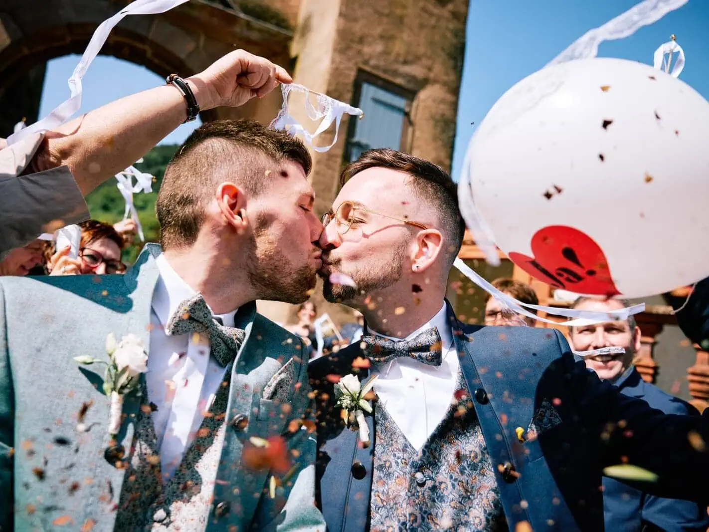Unter einem Regen aus Konfetti schreiten die Bräutigame aus der Trauungszeremonie, ein Kuss vereint sie. Umgeben von Hochzeitsgästen.