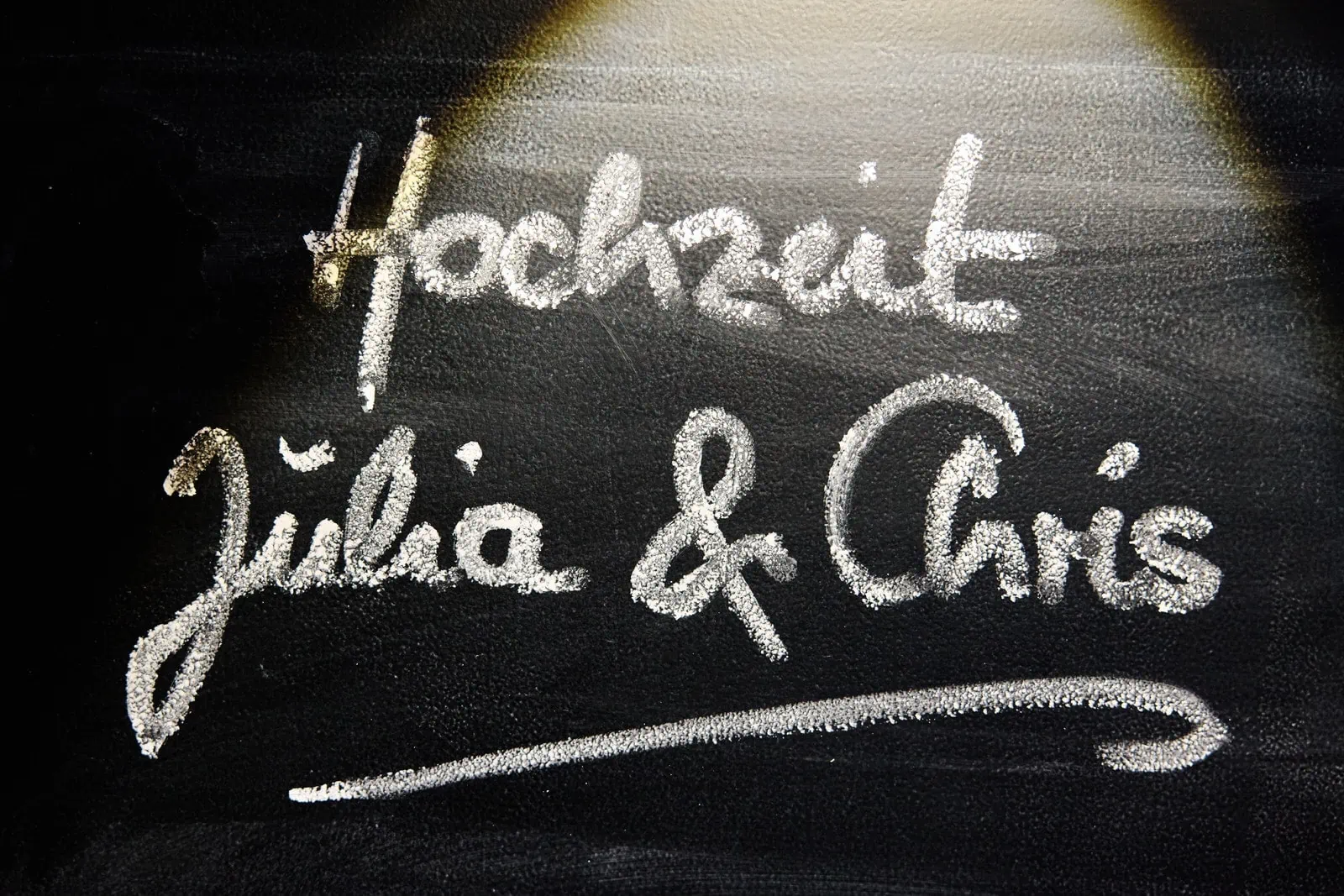 Eine Tafel auf der Hochzeit Julia und Chris mit Kreide geschrieben ist. Sie wird von einem Lichtkegel angestrahlt.