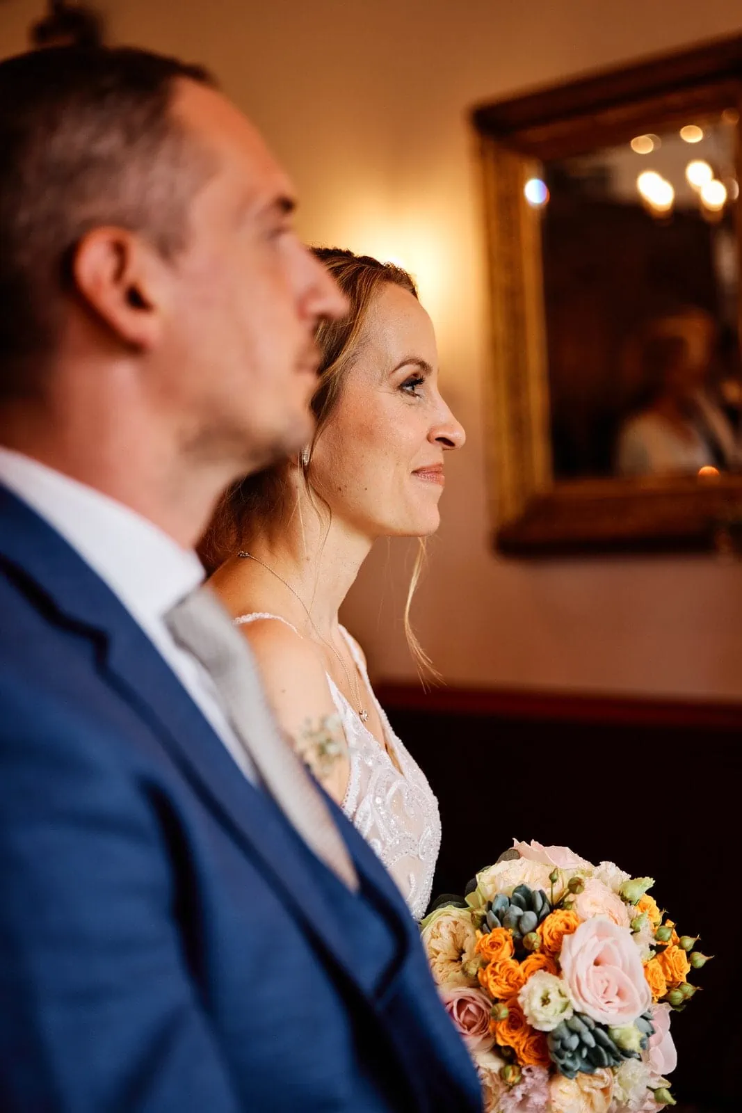 Die Braut ist im Fokus, der Bräutigam sitzt neben ihr und ist unscharf im Vordergrund zu sehen. Sie trägt ihren Brautstrauß und lächelt.