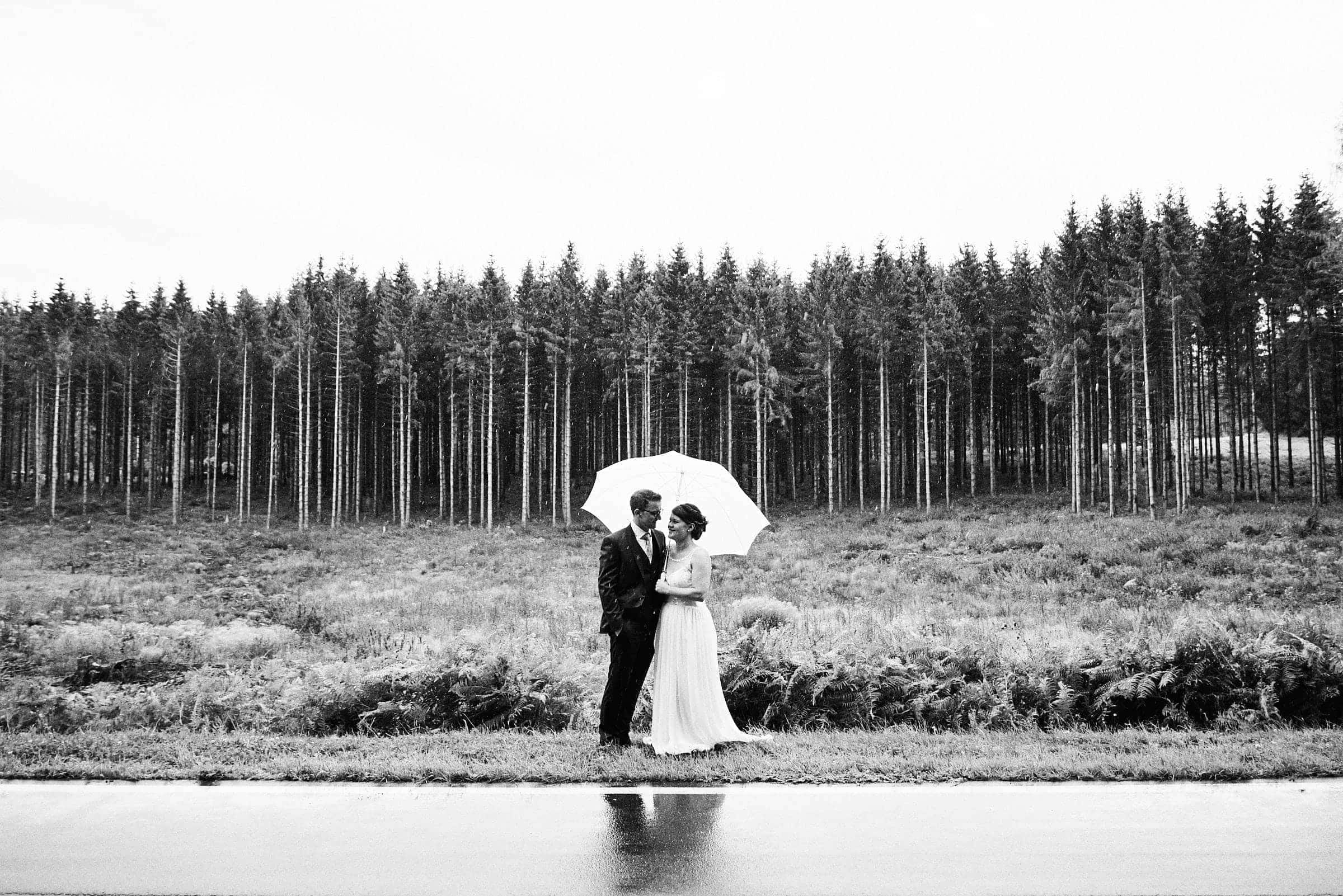 Brautpaar steht zusammen unter einem weißen Regenschirm am Fluss. Im Hintergrund erstreckt sich ein großer Nadelwald. Die Aufnahme ist in schwarzweiß.