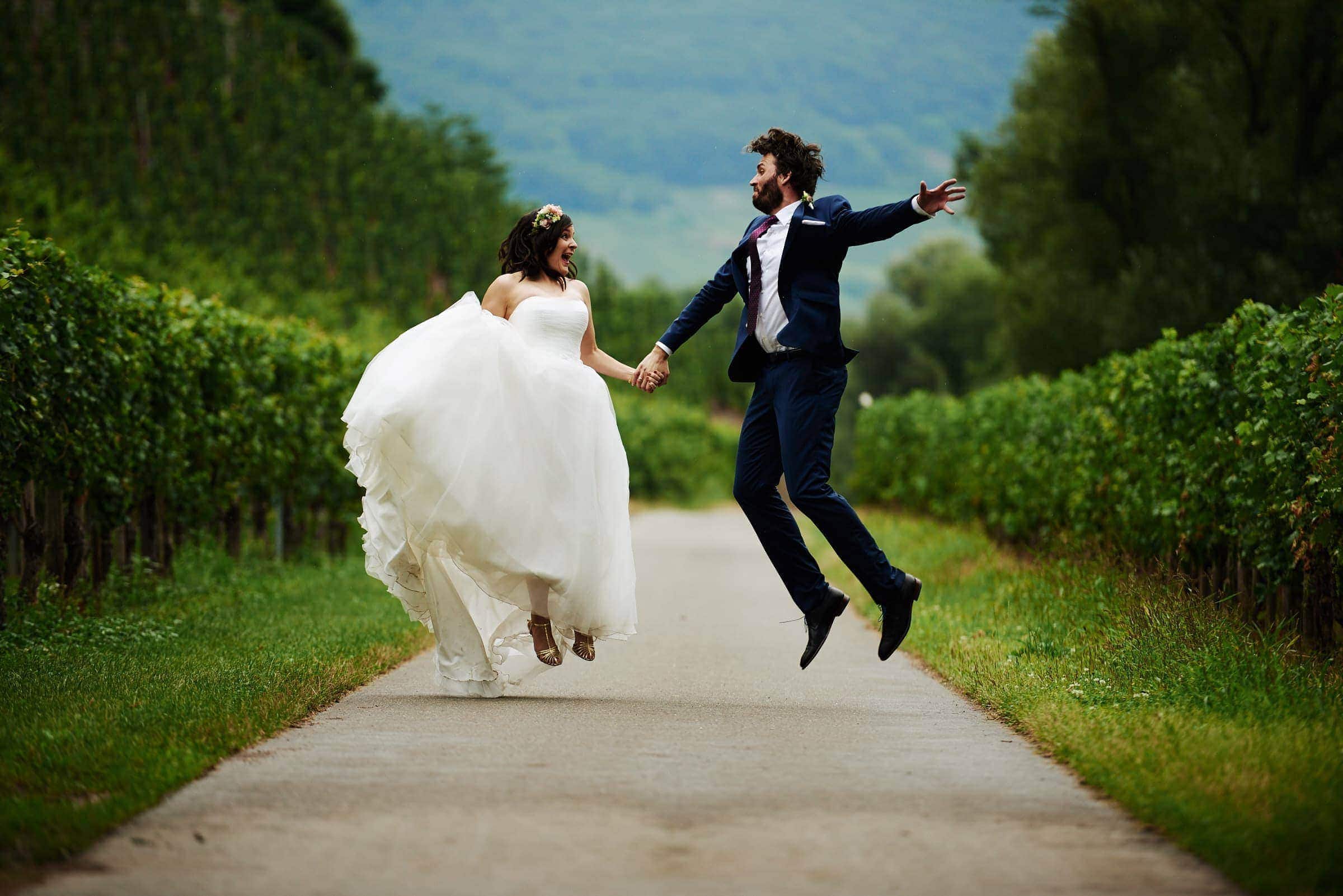 Ein Brautpaar auf einem Landweg zwischen Weinbergen. Sie springen energisch in die Luft und halten dabei ihre Hände.