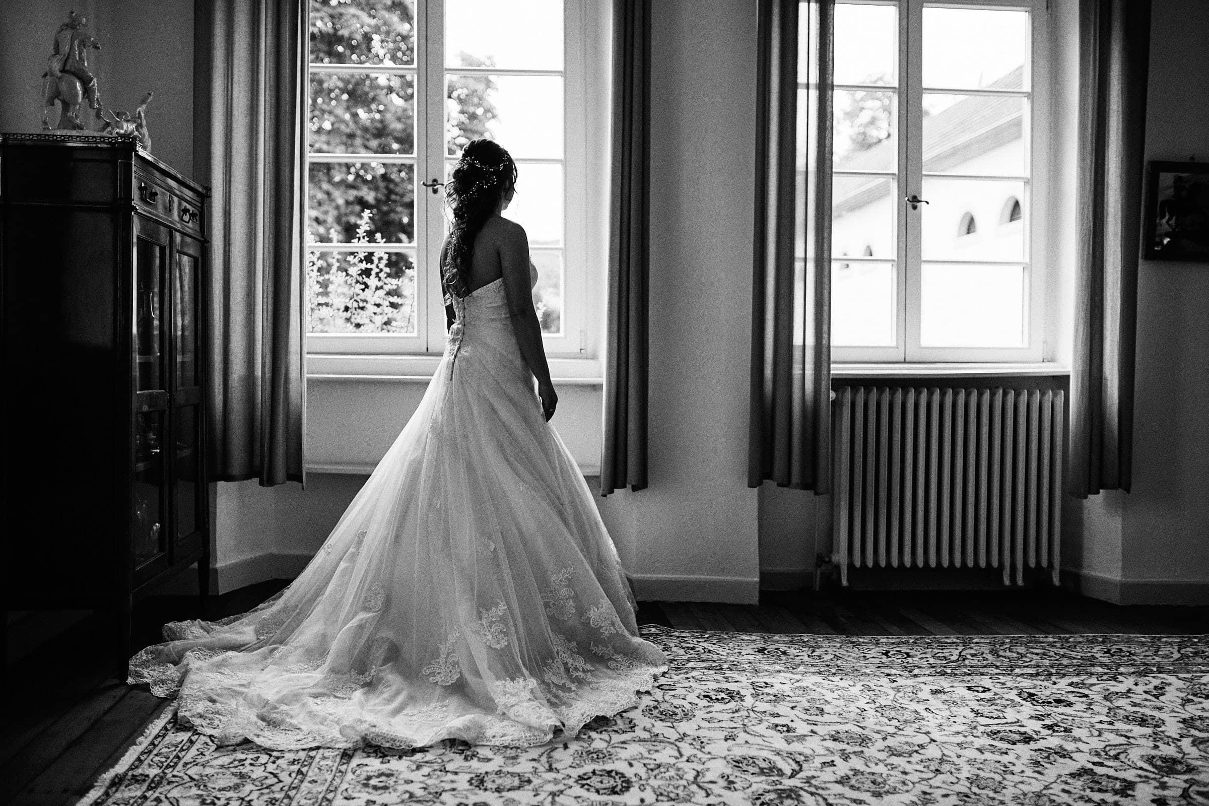 Schwarzweiße romantische Fotografie, die Braut steht links am Fenster und schaut in ihre Zukunft.