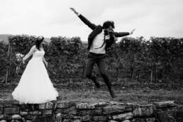 Bräutigam springt mit Eleganz von der Mauer und streckt die Arme aus. Die Braut steht daneben und macht einen sehr überraschten Blick. Die Aufnahme ist in schwarzweiß.